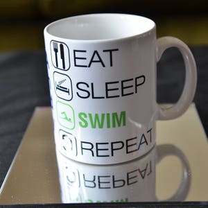 May include: White ceramic mug with the words "EAT SLEEP SWIM REPEAT" in black and green. The mug is on a mirrored surface, reflecting the text. The mug has a curved handle.