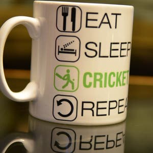 May include: White ceramic mug with black and green text and icons. The text reads "Eat, Sleep, Cricket, Repeat".