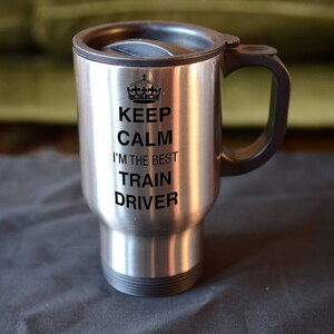 May include: Stainless steel travel mug with a black lid and handle. The mug features a black and white graphic that reads "Keep Calm I'm the Best Train Driver".