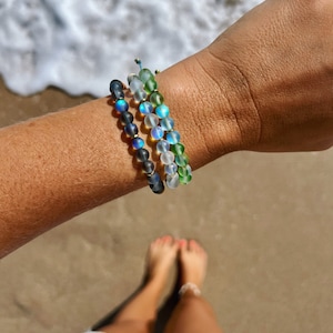May include: Three layered bracelets with iridescent glass beads in shades of blue, green, and white. The bracelets are worn on a person's wrist.