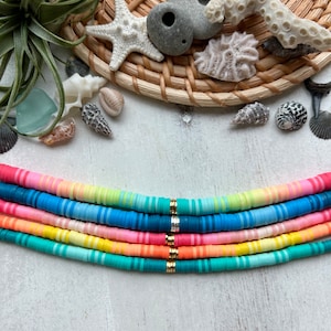 Peut inclure: Un collier de perles plates et rondes de différentes couleurs, disposées en dégradé de rose à turquoise.