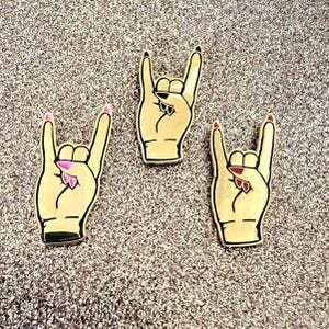 May include: Three gold-colored metal pins shaped like a hand making the rock and roll hand gesture. Each pin has a different color nail polish on the fingers: pink, black, and red.