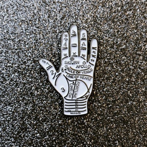 May include: White enamel pin in the shape of a palm, featuring black line drawings of palmistry symbols and lines. The pin includes text such as "Saturn," "Apollo," and "Line of Heart." The pin is set against a glittery background.