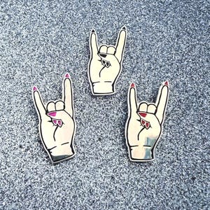 May include: Three silver metal pins with a hand making the rock and roll hand gesture. The hand is white with pink nail polish.