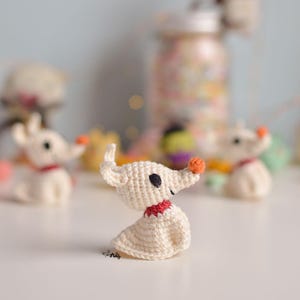 Pode incluir: Estatueta de cão branco de crochê com coleira vermelha e nariz laranja. O cão tem olhos pretos e está sentado em posição vertical. Outras figuras semelhantes estão no fundo. As figuras são inspiradas no personagem Zero de O Estranho Mundo de Jack.