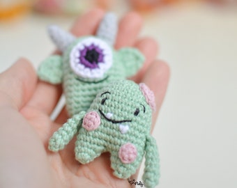 May include: Two crocheted green monster toys. One monster has a single large purple eye and two small horns. The other monster has two small pink ears and a toothy smile.