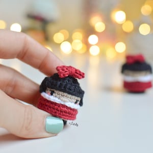 May include: Two small crocheted dolls with red and white clothing and black hair. The doll on the left is wearing a red bow. The dolls are sitting on a white surface.
