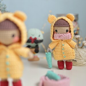 May include: Two crocheted dolls wearing yellow raincoats with brown hair and pink masks. One doll is holding a teal umbrella. The dolls are standing on a white surface.
