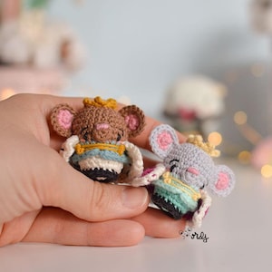 Puede incluir: Dos pequeñas figuras de animales de crochet que llevan coronas y túnicas. La figura marrón tiene la nariz y las orejas rosas, y la figura gris tiene la nariz y las orejas rosas. Ambas figuras sostienen un pequeño objeto de crochet en sus manos.