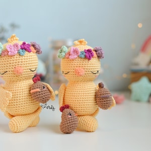 Puede incluir: Dos pollitos amarillos de crochet con coronas de flores y pollitos marrones en sus brazos. Los pollitos están de pie sobre una superficie blanca.