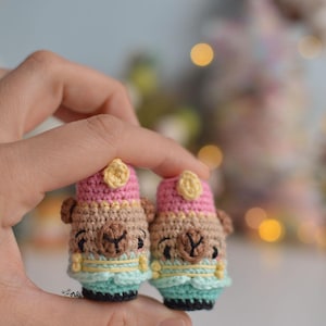 Puede incluir: Dos pequeñas figuras de llama de ganchillo hechas a mano. Cada llama tiene un sombrero rosa y beige con un acento amarillo, una cara marrón y un ribete turquesa y amarillo. Las llamas se sostienen entre los dedos.