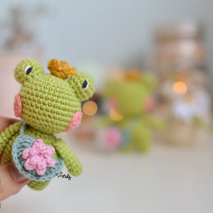 May include: A small green crocheted frog with a pink flower on its chest and a yellow crown on its head. The frog is holding a small blue crocheted bag with a pink flower on it.
