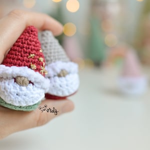May include: Two crocheted gnomes, one with a red hat and green beard, the other with a grey hat and white beard. Both gnomes have white fur trim and gold accents.