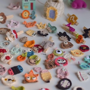 May include: A collection of small, colorful, crocheted appliques featuring various shapes and characters, including animals, food items, and abstract forms. Many of these miniature textile pieces are attached to wooden clothespins, scattered across a surface.