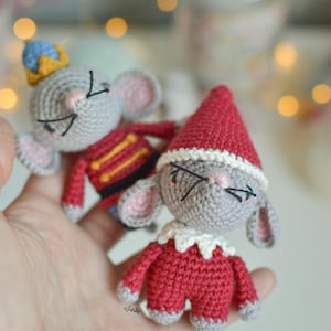 Könnte beinhalten: Zwei gehäkelte Mausschmusetiere, eines als Nussknacker verkleidet mit einer rot-goldenen Uniform und einer blauen Krone, das andere als Elf mit einer roten Mütze und einem rot-weißen Outfit.