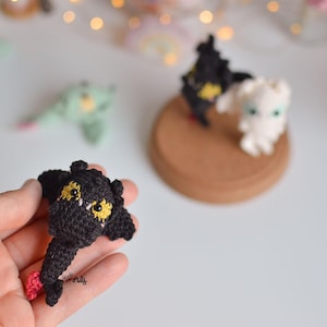 Puede incluir: Figuras de dragones de ganchillo en negro, blanco y verde. El dragón negro se sostiene en una mano, mostrando ojos amarillos y una lengua rosa. Otros dragones están sobre un posavasos de madera. Las figuras son pequeñas y detalladas.