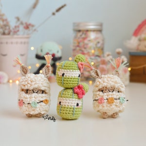 Può includere: Tre giocattoli amigurumi all'uncinetto: un cactus verde con fiori rosa e due lama bianchi con accenti colorati. I giocattoli sono su una superficie bianca.