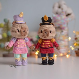 Puede incluir: Dos muñecos de oso de ganchillo hechos a mano vestidos de cascanueces. Uno lleva un atuendo rosa y morado con un sombrero a juego, el otro un uniforme rojo y dorado con un sombrero negro. Ambos tienen cuerpos marrones y zapatos negros.