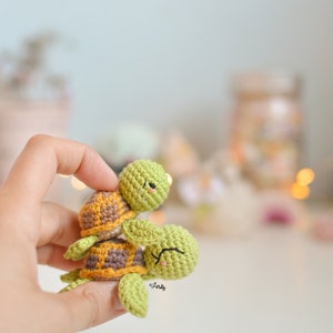 Puede incluir: Dos tortugas de crochet verde con caparazones marrones y amarillos. La tortuga más pequeña está montada sobre la más grande.