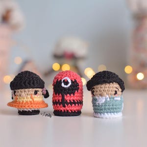 Puede incluir: Tres figuras de crochet, una con un atuendo rojo y negro, otra con un atuendo azul y blanco y otra con un atuendo naranja y amarillo.