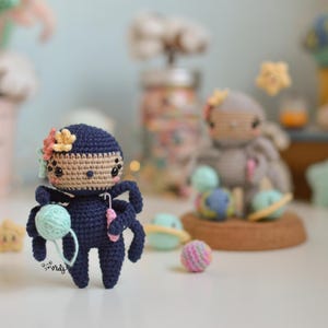 Puede incluir: Un juguete de araña amigurumi de crochet con un cuerpo azul, una cabeza marrón y detalles de flores rosas y amarillas. La araña sostiene una bola de hilo y un gancho de crochet.