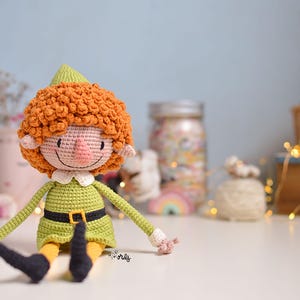 Puede incluir: Una muñeca de elfo de crochet con pelo rizado naranja, un vestido verde y un cinturón marrón. El elfo está sentado sobre una superficie blanca.