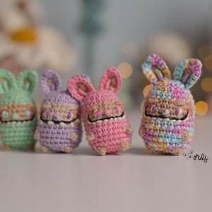 Peut inclure: Quatre petits lapins crochetés aux couleurs pastel : vert, violet, rose et multicolore. Chaque lapin a de grandes oreilles, un petit visage avec des détails noirs et un corps arrondi. Les lapins sont disposés en rangée.