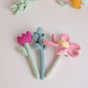 Puede incluir: Tres juguetes de flores de crochet con tallos. Las flores son rosas, azules y verdes. Los tallos son verdes, azules y beige.