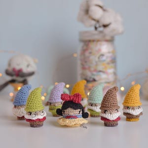 May include: A group of seven crocheted gnomes and a crocheted Snow White doll. The gnomes are all different colours, including blue, green, purple, brown, and yellow. Snow White is wearing a yellow dress with a red bow in her hair.