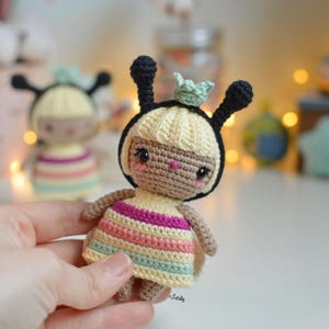 May include: A small crocheted doll with a striped dress, black antennae, and a green crown. The doll has brown eyes and a light brown complexion.