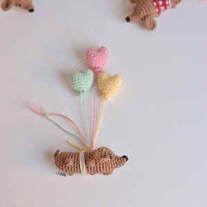 Può includere: Un bassotto di uncinetto marrone che tiene tre palloncini a forma di cuore. I palloncini sono rosa, verde e giallo.