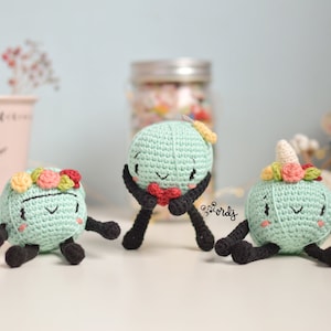 Puede incluir: Tres muñecas de ganchillo hechas a mano en color verde claro, cada una con una corona floral única. Una muñeca tiene un corazón, otra un cuerno de unicornio. Tienen brazos y piernas negros y caras sonrientes. Las muñecas están sobre una superficie blanca.