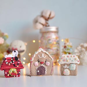Puede incluir: Tres casas de jengibre de ganchillo, cada una con detalles únicos. Una es roja con ribetes blancos y un muñeco de nieve en la parte superior, otra es marrón con una puerta blanca y una cara sonriente, y la última es beige con decoraciones de caramelo de colores.