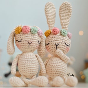 May include: Two crocheted bunny toys with closed eyes and flower crowns. The bunnies are cream colored with pink, green, and yellow flowers.