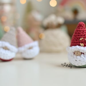 Op de afbeelding: Een rode gehaakte gnome met een witte baard en gouden glitteraccenten. De gnome zit op een wit oppervlak.