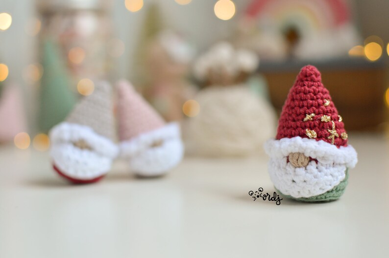 Op de afbeelding: Een rode gehaakte gnome met een witte baard en gouden glitteraccenten. De gnome zit op een wit oppervlak.