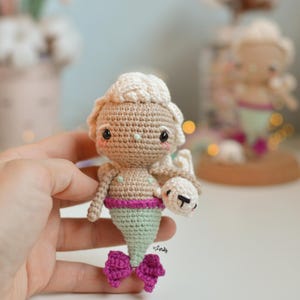 May include: Crocheted mermaid doll with light brown skin, a light green tail, and a pink top and bow. The doll has a light brown hair and is holding a small white bear. The doll is handmade.