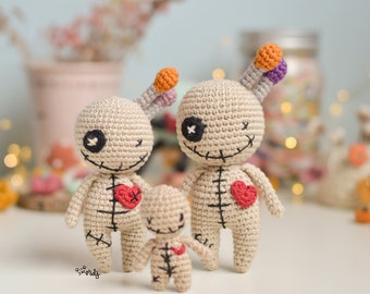 May include: Three crocheted dolls in a family arrangement. The dolls have tan bodies, black stitched details, and red heart accents. The ears are striped with orange, purple, and gray. The dolls have stitched smiles and one eye is a stitched X.