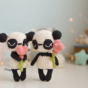 Puede incluir: Dos osos panda de crochet sosteniendo corazones rosas. Los pandas son blancos con orejas, ojos y pies negros. Están de pie sobre una superficie blanca.