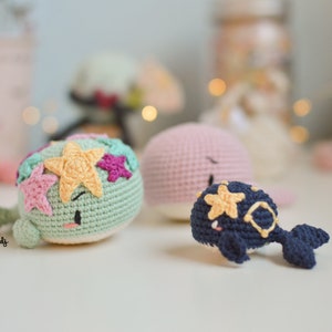 Puede incluir: Tres peluches de ballena de crochet. Una es verde con estrellas rosas, amarillas y moradas. Otra es rosa. La última es azul con estrellas amarillas.