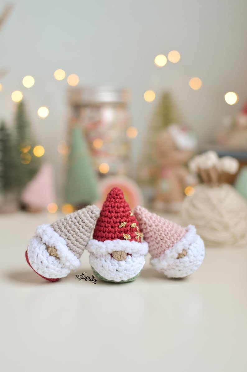 Op de afbeelding: Drie gehaakte gnomen met verschillende gekleurde mutsen. De gnome links heeft een beige muts, de middelste gnome heeft een rode muts met gouden glitter en de gnome rechts heeft een roze muts.