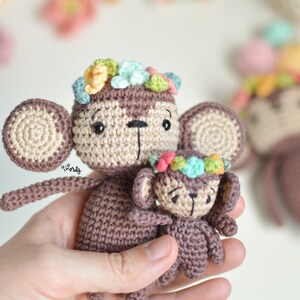 Può includere: Due giocattoli di scimmia all'uncinetto con corone di fiori. La scimmia più grande tiene la scimmia più piccola. Entrambe le scimmie sono marroni con volti beige e occhi neri.