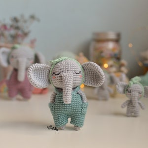 Puede incluir: Tres juguetes de elefante de crochet. El elefante del centro lleva una diadema verde y un mono verde y gris. Los otros dos elefantes llevan atuendos rosa y grises.