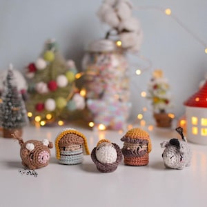 Puede incluir: Una colección de cinco figuras de nacimiento hechas a mano con ganchillo, que incluyen un cerdo, dos figuras con sombreros, una oveja y una vaca. Las figuras son marrones, beige y grises, con detalles blancos. Decoraciones navideñas en el fondo.