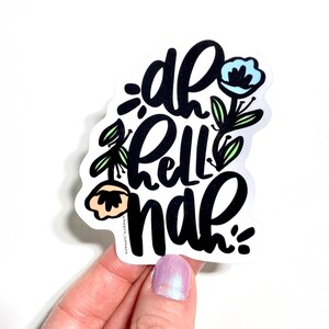 Puede incluir: Una pegatina blanca con la frase "oh hell nah" en escritura negra, adornada con ilustraciones florales en azul claro, melocotón y verde. La pegatina está sujeta por una mano con esmalte de uñas lila claro.