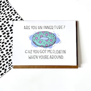May include: A greeting card with a light blue background and a cartoon illustration of a green and pink inner tube with white polka dots. The text on the card reads "Are you an inner tube? Cuz you got me floatin' when you're around."