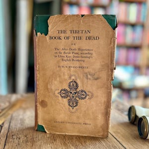 May include: A vintage book titled "The Tibetan Book of the Dead" with a green spine and a brown cover. The cover features a black and white illustration of a Tibetan symbol.