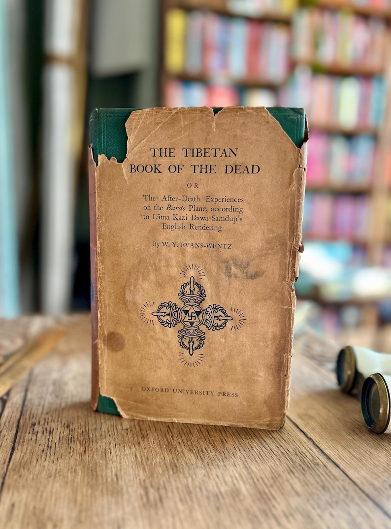May include: A vintage book titled "The Tibetan Book of the Dead" with a green spine and a brown cover. The cover features a black and white illustration of a Tibetan symbol.