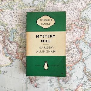 Puede incluir: Una edición vintage de Penguin Books de "Mystery Mile" de Margery Allingham. El libro tiene una cubierta verde con texto blanco y un logotipo de pingüino. El libro está sobre un mapa del mundo antiguo.