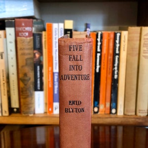 Puede incluir: Un libro de tapa dura marrón con el título "Five Fall Into Adventure" y el autor "Enid Blyton" en el lomo. El libro está en una estantería con otros libros.